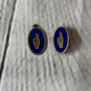 Op de afbeelding: Twee ovale hangers met een blauwe achtergrond en een gouden handgebaar. E&eacute;n heeft een lus, de andere is een speld. Beide hebben een gouden rand. Waarschijnlijk vintage.