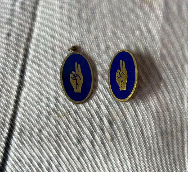 Op de afbeelding: Twee ovale hangers met een blauwe achtergrond en een gouden handgebaar. E&eacute;n heeft een lus, de andere is een speld. Beide hebben een gouden rand. Waarschijnlijk vintage.