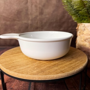 May include: A white ceramic bowl with a handle, set on a round wooden stand with black metal legs. The bowl has a simple, clean design. The background is a brown, textured surface.