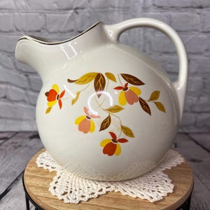 May include: Cream-colored ceramic pitcher with a rounded body and a handle. The pitcher is decorated with a floral pattern in shades of yellow, orange, and brown. The rim is trimmed with a gold-colored band. The pitcher is sitting on a small wooden stand.