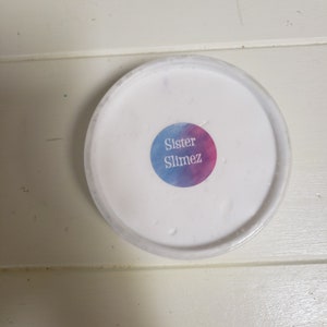 May include: A white container of slime with a clear lid. A round sticker on the lid reads "Sister Slimez" in white text over a blue and pink watercolor background. The container sits on a white wooden surface.