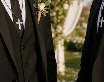May include: Two black suits with matching black vests, white dress shirts, and black ties. Each suit features a silver cross pendant on a black cord necklace and a white rose boutonniere with green leaves wrapped in twine on the left lapel.