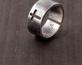May include: A silver-toned metal ring with a brushed finish featuring a cutout cross symbol on the band. The interior of the ring is engraved with the letters KEA. The band has an open design, showing a gap on the side of the ring structure.