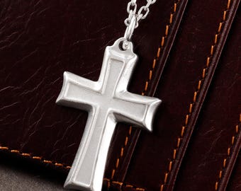 May include: A silver-toned cross pendant suspended from a matching silver-toned metal chain. The cross features a smooth, matte-finished center panel bordered by raised, polished edges, creating a distinct layered appearance in its design.
