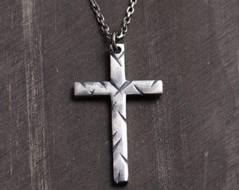 May include: A silver-toned metal cross pendant suspended from a matching thin link chain. The cross features a brushed, matte finish with distinct, short, diagonal etched marks across its surface, creating a textured and weathered appearance.