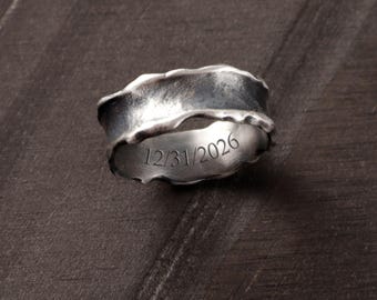 May include: A silver ring with an irregular, textured outer surface and a smooth inner band. The interior of the ring is engraved with the date 12/31/2026 in a clear, serif font. The metal has a darkened, oxidized finish throughout the piece.