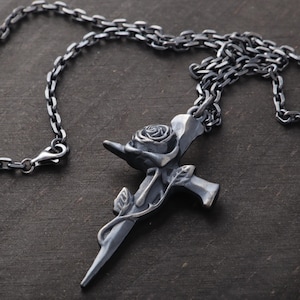 May include: A silver-toned pendant necklace featuring a cross design with a rose and leaves. The pendant is attached to a chain with a lobster clasp. The necklace is displayed on a dark wooden surface.