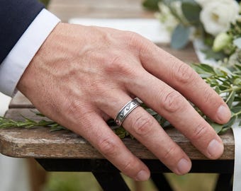 May include: A silver ring with a wave pattern design. The ring is on a person's finger, with a dark blue suit and white shirt sleeve visible. The background includes greenery and a wooden surface.