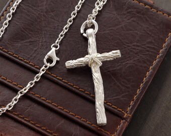 May include: A silver-colored cross pendant necklace with a textured, wood-like design. The cross is suspended from a delicate chain with a lobster clasp. The necklace rests on a brown leather wallet.
