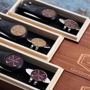 May include: Three wooden boxes with personalized golf divot tools and ball markers. Each box has a different name engraved on it: "Derrick", "Lucca", and "Logan". The boxes are made of light brown wood and have a dark brown stain. The boxes are arranged in a row, with the "Derrick" box on the left, the "Lucca" box in the middle, and the "Logan" box on the right.