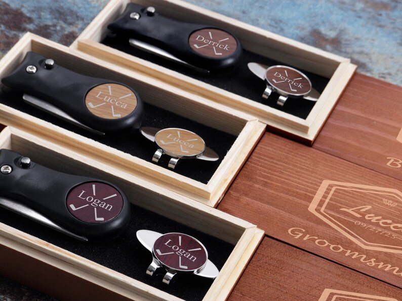 May include: Three wooden boxes with personalized golf divot tools and ball markers. Each box has a different name engraved on it: "Derrick", "Lucca", and "Logan". The boxes are made of light brown wood and have a dark brown stain. The boxes are arranged in a row, with the "Derrick" box on the left, the "Lucca" box in the middle, and the "Logan" box on the right.