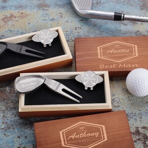 May include: Two wooden boxes with engraved names, dates, and the words "Best Man". Each box contains a golf ball retriever and a golf ball marker with crossed golf clubs. One box is brown and the other is light wood. A white golf ball is sitting on the table next to the boxes.