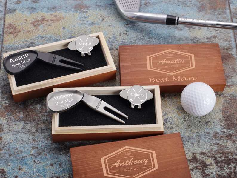 May include: Two wooden boxes with engraved names, dates, and the words "Best Man". Each box contains a golf ball retriever and a golf ball marker with crossed golf clubs. One box is brown and the other is light wood. A white golf ball is sitting on the table next to the boxes.
