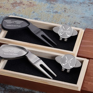 May include: Two personalized golf divot tools and hat clips in a wooden box. The divot tools are silver and black with engraved names and initials. The hat clips are silver with engraved initials and crossed golf clubs.
