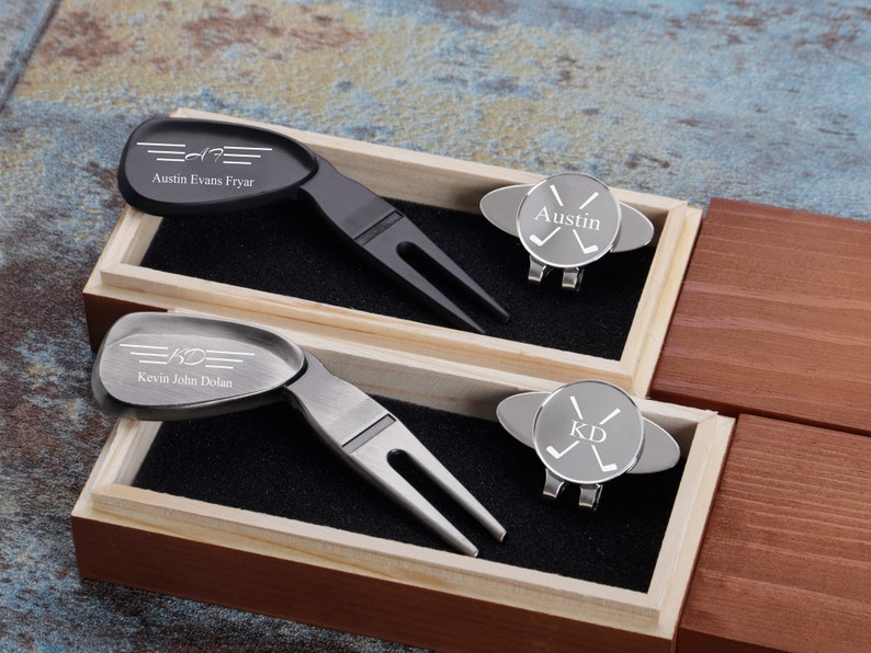 May include: Two personalized golf divot tools and hat clips in a wooden box. The divot tools are silver and black with engraved names and initials. The hat clips are silver with engraved initials and crossed golf clubs.