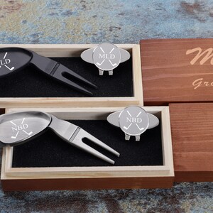 May include: Two wooden boxes with engraved lids. The lids are personalized with the names "MLD" and "NBD" and the date "11/18/2022". Each box contains a silver golf ball marker and a black or silver golf divot tool.