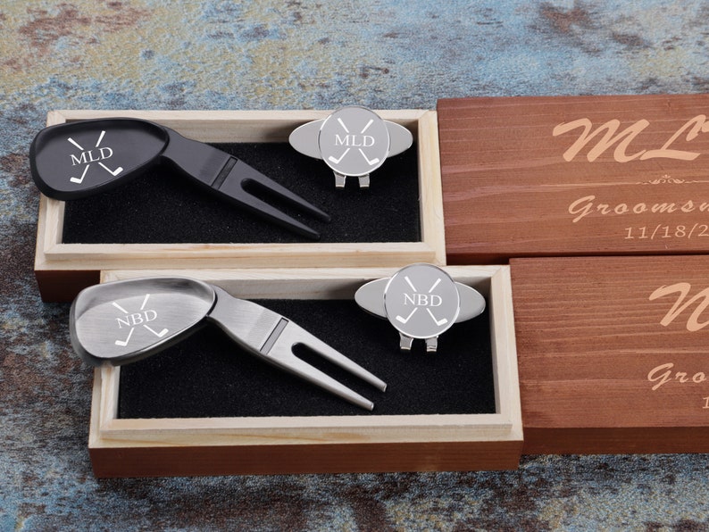 May include: Two wooden boxes with engraved lids. The lids are personalized with the names "MLD" and "NBD" and the date "11/18/2022". Each box contains a silver golf ball marker and a black or silver golf divot tool.