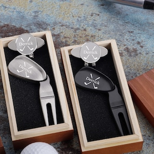 May include: Two wooden boxes with black velvet interiors, each containing a personalized golf ball marker and divot tool. The tools are engraved with the name "Derrick" and crossed golf clubs.