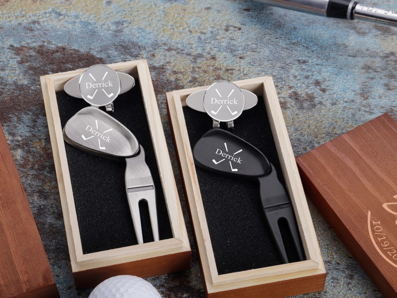 May include: Two wooden boxes with black velvet interiors, each containing a personalized golf ball marker and divot tool. The tools are engraved with the name "Derrick" and crossed golf clubs.