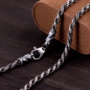 May include: A silver-toned rope chain necklace featuring a dark, oxidised finish. The chain is constructed from twisted metal links and includes a secure lobster claw clasp mechanism, flanked by two decorative, spiralled cylindrical end caps.