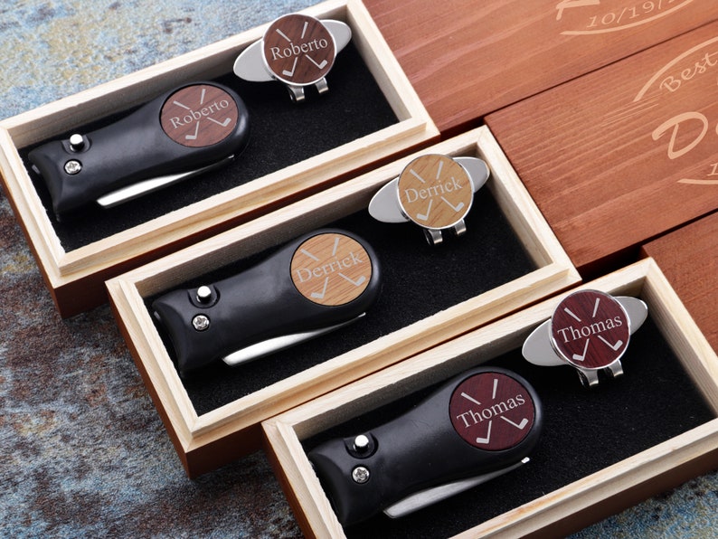 May include: Four wooden boxes with black golf tool sets inside. Each box has a personalized golf ball marker with a name and crossed golf clubs. The names are Roberto, Derrick, Thomas, and Thomas.