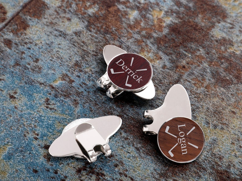 May include: Two silver golf ball markers with wooden inserts. The inserts are engraved with the names "Derrick" and "Logan" and a golf club graphic.