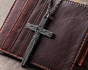 May include: A dark gray metal crucifix pendant featuring a detailed figure of Jesus, suspended from a matching dark gray metal chain. The cross has a textured, sunburst-like design radiating from the center, resting against a brown leather wallet.