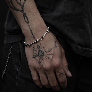 May include: A silver bracelet with a bone-shaped design. The bracelet is worn on a wrist with tattoos. The jewellery appears to be made of metal and has a unique, edgy aesthetic. The bracelet is a stylish accessory.