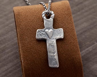 May include: A silver-toned metal cross pendant featuring a textured, hammered surface and a small raised heart detail at the center. The cross hangs from a matching silver-toned metal chain, with a twisted metal loop connecting the two pieces.