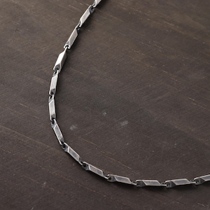 May include: A silver-colored necklace with a geometric bar link design. The necklace is displayed on a dark brown wooden surface. The links are rectangular with angled edges, creating a modern, industrial aesthetic.