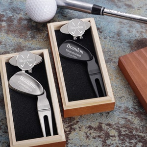 May include: Two personalized golf divot tools in wooden boxes. One tool is silver with a black divot tool and the other is black with a silver divot tool. Both tools have a golf ball marker with the name "Brandon" and the text "Groomsman 12.19.2021".