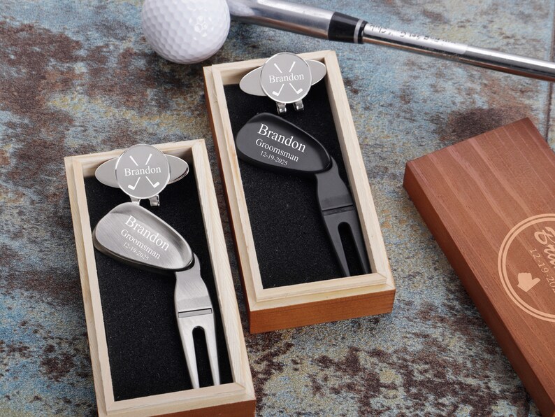 May include: Two personalized golf divot tools in wooden boxes. One tool is silver with a black divot tool and the other is black with a silver divot tool. Both tools have a golf ball marker with the name "Brandon" and the text "Groomsman 12.19.2021".