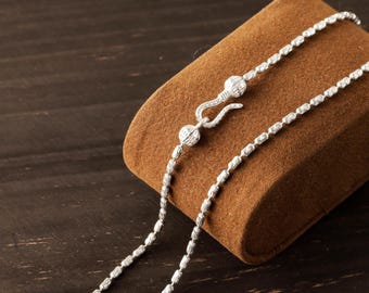 May include: A silver chain necklace featuring a series of small, textured, cylindrical links. The necklace is secured with a hook-and-eye style clasp, which is flanked by two small, round, decorative beads with a hammered finish.