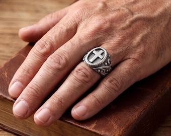 May include: A silver-toned metal ring featuring an oval face with a raised cross symbol in the center. The band and the setting are decorated with intricate, swirling scrollwork patterns and a beaded border surrounding the central cross design.
