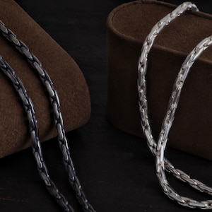 May include: Two silver-colored chain necklaces with a textured, interlocking design. One necklace is a darker, oxidized silver, while the other is bright silver. The chains are displayed on a brown, velvet-like surface.