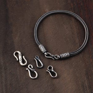 May include: A silver-toned bracelet with a braided design and matching clasps. The bracelet is circular, with decorative clasps and several hook-style clasps scattered around it. The background is a dark brown wooden surface.