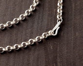 May include: A silver rolo chain necklace featuring a series of interconnected round links and a hook-style clasp. The metal has a matte finish, and the chain is arranged in a curved line, showcasing the uniform circular shape of each individual link.