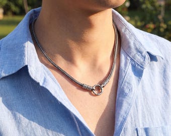 May include: A silver-toned chain necklace featuring a woven, textured wheat-style link design. The necklace includes a prominent circular clasp mechanism at the front, which connects the two ends of the chain with detailed, engraved metal accents.