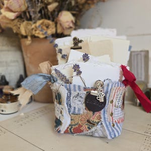 Puede incluir: Una caja de tela con diseño de patchwork, llena de papel blanco y lavanda seca. La caja presenta tela a rayas azules y blancas, estampados florales y una etiqueta de cuero que dice "sketch". Una cinta roja y clips metálicos añaden detalles.