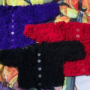 May include: Three crocheted baby sweaters in purple, red, and black. Each sweater has a button fastening.
