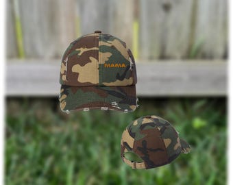 May include: Camouflage baseball cap with the word "mama" embroidered in orange thread. The cap features a distressed, worn look with frayed edges. The hat is in shades of brown, green, and tan.