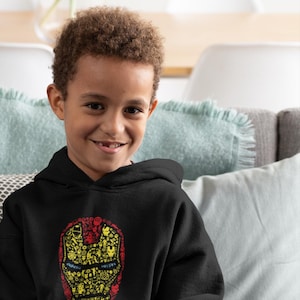 Puede incluir: Una persona joven con una sudadera con capucha negra con un gráfico de la cara de Iron Man en amarillo y rojo hecho de imágenes más pequeñas. La persona está sentada en un sofá y sostiene un mando de videojuego gris.