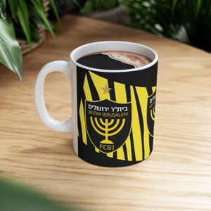 Pode incluir: Uma caneca de cerâmica branca com um design listrado preto e amarelo. A caneca apresenta o logotipo do clube de futebol israelense Beitar Jerusalém, que inclui um candelabro e o texto "Beitar Jerusalém" em hebraico. A caneca também tem o texto "FCBJ" na parte inferior.
