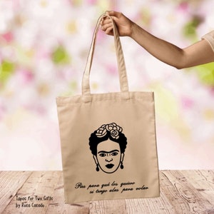May include: A beige canvas tote bag with black handles. The bag features a black and white illustration of Frida Kahlo with the quote "Pies para qué los quiero si tengo alas para volar".