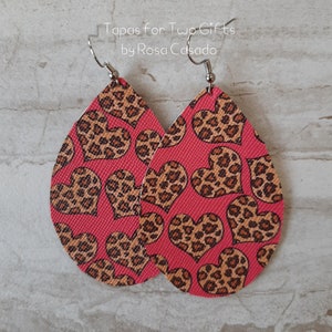 May include: A pair of pink teardrop-shaped earrings with a leopard print heart pattern. The earrings are made of leather and have silver hooks.