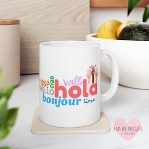 May include: White ceramic mug with the word "hold" in red, with "hello", "bonjour", "hola", "ciao", "你好", and "مرحبا" in various colors. The mug also features a graphic of hands in different skin tones.