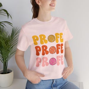 May include: A light pink short-sleeve t-shirt featuring the word PROFE repeated three times in a stacked, wavy font. Each line includes a circular concha bread graphic in place of the letter O, colored in yellow, orange, and pink respectively.