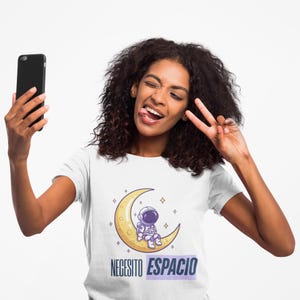 May include: White t-shirt featuring a cartoon astronaut seated on a yellow crescent moon. The shirt has the text "NECESITO ESPACIO" in blue and white. The person is holding a black smartphone and making a peace sign.
