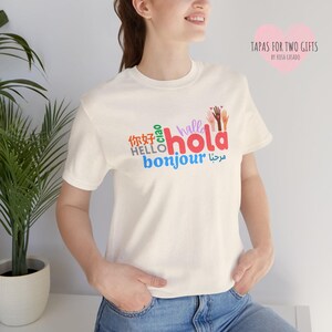 May include: Cream-colored t-shirt with the word "hola" in red, surrounded by "hello" in English, "bonjour" in blue, and other greetings in different languages. The shirt also features a graphic of colorful hands.
