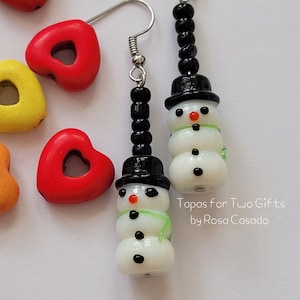 May include: A pair of snowman earrings with black beads, black hats, white snowman faces, and green scarves. The earrings are handmade and have a whimsical charm. "Tapas for Two Gifts by Rosa Casado" is written below the earrings.